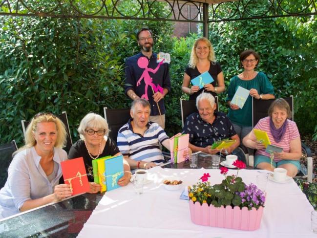 Großes Geschenkeauspacken im Garten von Haus Anrode (v.l.n.r.):  sitzend: Anja Kuypers (Initiatorin), Brigitte Fensterseifer, Dieter Heiß, Josef Winkels und Ingrid Haupt (Bewohner Haus Anrode) stehend: Jörg Marquardt (Bereichsleiter Haus Anrode) mit einem Bunten Menschen in der Hand, Barbara Holzhäuser und Christine Schierbaum (beides Betreuerinnen der Lebenshilfe)